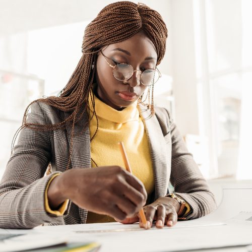 focused african american female architect in glasses holding pencil and working on project at desk